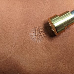 May include: A close-up of a brass leather stamping tool with a geometric design. The tool is being used to create an impression on a piece of brown leather.