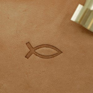 Könnte beinhalten: Nahaufnahme einer hellbraunen Lederoberfläche mit einem gestempelten Fischsymbol. Ein Metallwerkzeug befindet sich in der oberen rechten Ecke. Das Fischsymbol ist ein christliches Symbol, das oft in religiösem Kunsthandwerk und Lederarbeiten verwendet wird.