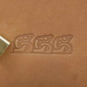 May include: Close-up of a leather stamping tool creating a repeating pattern of stylized bird or serpent-like figures on a tan leather surface. The tool is partially visible, showing a metallic finish. The stamped design is a series of interconnected shapes.
