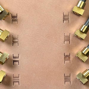 May include: Close-up of leather tooling stamps with various geometric designs. The stamps are made of brass and are arranged around the edges of a piece of tan leather. The leather has several stamped impressions of the designs.