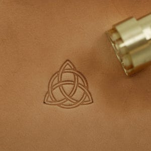 May include: A leather craft tool with a brass stamp is positioned near a stamped Celtic knot design. The knot is a series of interwoven lines, and the leather has a light brown color.