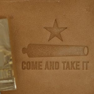 May include: Close-up of a tan leather surface with an engraved design. The design includes a star, a cannon, and the words "COME AND TAKE IT". The leather has a textured appearance.