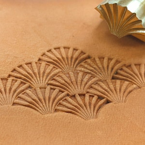 Puede incluir: Un primer plano de una herramienta de estampado de cuero con un diseño en forma de abanico. La herramienta está hecha de latón y tiene un borde afilado. La herramienta se utiliza para crear un patrón en un trozo de cuero.