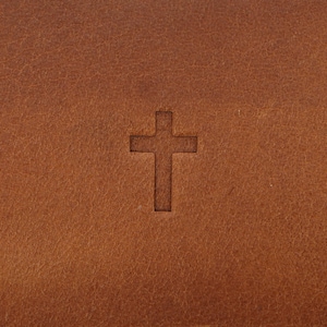 May include: Close-up of a brown leather surface with a debossed cross symbol. The leather has a textured appearance and a warm, earthy tone. The cross is centered, adding a religious or spiritual element to the design.