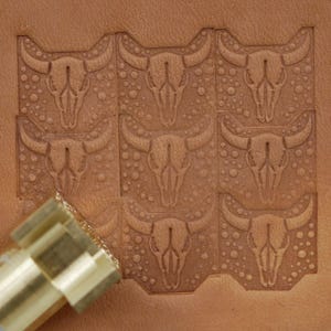 May include: A leather stamping tool in use, creating a repeating pattern of bull skulls. The tool is gold-coloured, and the leather is light brown. The design includes small, circular details around the skulls.