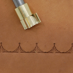 May include: A close-up of a leather stamping tool with a decorative fan-shaped design. The tool is made of brass and is positioned above a piece of brown leather that has been stamped with the same fan pattern.