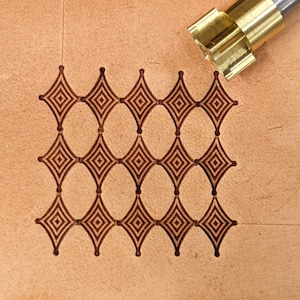 Op de afbeelding: Een close-up van een lederbewerkingstool die wordt gebruikt om een ​​herhalend patroon van ruiten en ovalen te maken. De tool is gemaakt van messing en heeft een scherpe, puntige punt. De tool wordt gebruikt om een ​​decoratief ontwerp op leer te maken.