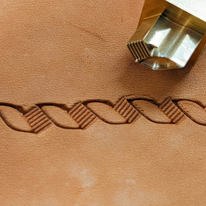 May include: A close-up of a leather stamping tool with a braided design. The tool is made of brass and has a textured surface. The tool is being used to create a braided design on a piece of leather.