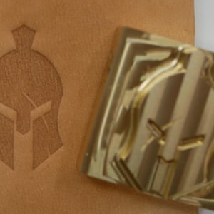 May include: A tan leather surface with a Spartan helmet design is shown next to a gold-colored metal stamp. The stamp is square and features a raised Spartan helmet design. The leather has a debossed helmet image.