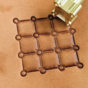 May include: A close-up of a leather stamping tool with a grid pattern. The tool is made of brass and has a square shape. The tool is being used to create a grid pattern on a piece of leather.