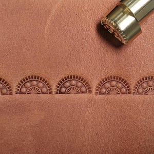 May include: Close-up of a leather surface with five embossed designs of a fan shape with a circle pattern. A brass tool is visible in the top right corner of the image.