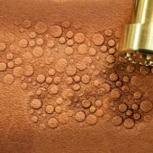May include: A close-up of a leather surface with a pattern of embossed circles created by a brass tool. The tool is visible in the top right corner of the image.