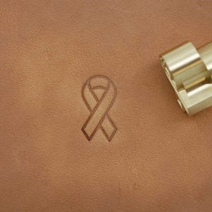 May include: A leather craft with a ribbon design stamped into the surface. The ribbon is a symbol of awareness. A brass branding iron is in the upper right corner. The leather is a light brown color.