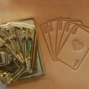 May include: A brass playing card stamp next to an embossed leather surface. The embossed design shows a hand of cards: 10 of hearts, Jack of hearts, Queen of hearts, King of hearts, and Ace of hearts.