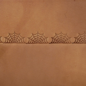 May include: Close-up of a brown leather surface with a repeating spiderweb pattern. The spiderweb design is stamped into the leather, creating a textured effect. A tool is visible in the upper left corner, suggesting the process of creating the design.