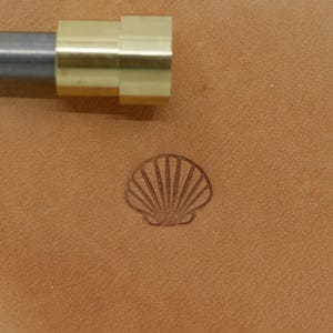 May include: A metal and brass leather stamping tool with a seashell design. The tool is silver and gold in color. The seashell stamp is pressed into a tan leather surface.