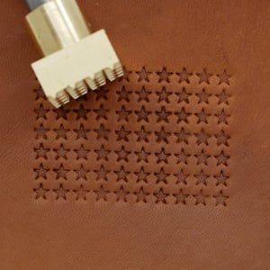 May include: A close-up of a brown leather surface with a pattern of stars stamped into it. A brass tool is positioned at the top, ready to stamp more stars. The stars are arranged in a flag-like pattern.