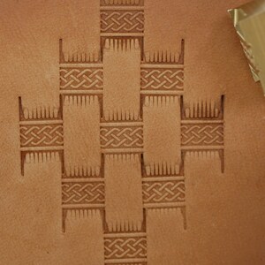 May include: Close-up of a tan leather surface with a geometric pattern of Celtic knots and vertical lines. A metal tool is in the upper right corner. The design is likely for leathercraft.