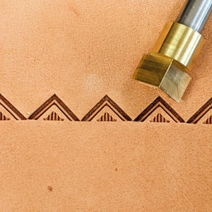 May include: A close-up of a leather stamping tool with a brass head and a wooden handle. The tool is being used to create a pattern of triangles on a piece of leather.