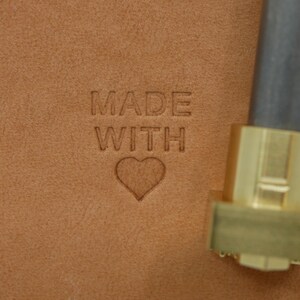 May include: Close-up of tan leather with the words "MADE WITH" and a heart symbol embossed into the surface. A metal tool with a brass head is in the right corner, suggesting a crafting process.
