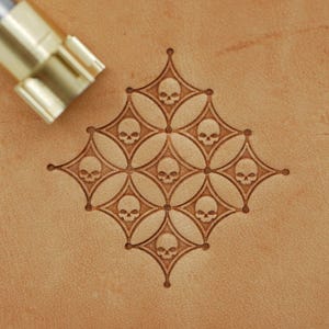 Op de afbeelding: Een leerstempelgereedschap naast een patroon van schedels. Het patroon bestaat uit herhalende ruitvormen met een schedel in elk. De tool is goudkleurig en heeft een metalen handvat.