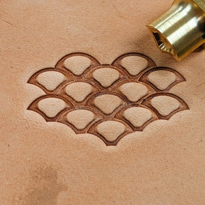 May include: A close-up of a leather craft tool with a brass handle and a design of overlapping scales. The tool is used to create a decorative pattern on leather.