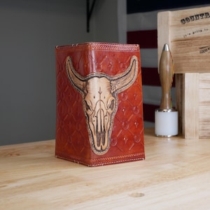 May include: A tall, rectangular, reddish-brown leather wallet featuring a hand-tooled steer skull design on the front. The leather surface has a repeating embossed geometric pattern and visible stitching along the edges, showcasing a detailed, natural finish.