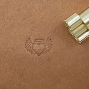 May include: A heart with wings and a halo is stamped into tan leather. A brass and silver metal stamp tool is in the upper right corner. The heart design is a common symbol used in leathercrafting and other crafts.