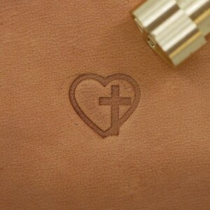 May include: A heart-shaped design with a cross inside, embossed on a tan leather surface. A metal tool is in the upper right corner. The heart and cross are a single continuous line, creating a religious symbol.