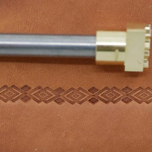 May include: A metal leather stamping tool with a geometric design. The tool has a silver handle and a brass stamping head. The stamped design is a repeating pattern of diamonds and squares on a brown leather surface.