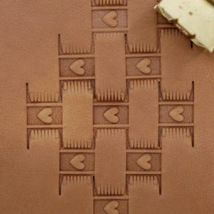May include: Close-up of a tan leather surface with a repeating pattern of hearts and rectangular shapes. The design is embossed into the leather. A small, light-colored tool rests in the upper right corner.