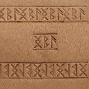 May include: A close-up of a piece of brown leather with a repeating pattern of Viking runes carved into it. The runes are arranged in a horizontal line at the top and bottom of the image, with a single square of runes in the center.