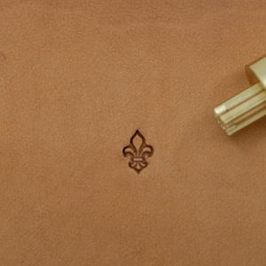 May include: A close-up of a tan leather surface with a fleur-de-lis stamp and a brass tool. The fleur-de-lis is stamped into the leather. The brass tool is in the upper right corner.