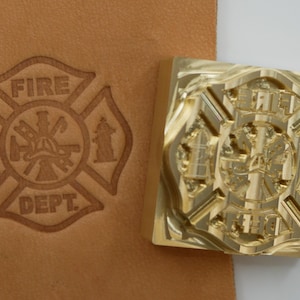 Könnte beinhalten: Ein Messingstempel mit dem Emblem der Feuerwehr neben einem geprägten Ledereindruck. Der Stempel ist ein detailliertes Kreuzdesign mit den Worten "FIRE DEPT.". Der Ledereindruck ist hellbraun.
