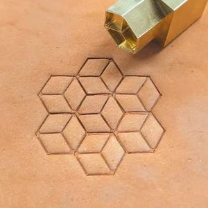 Könnte beinhalten: Ein Lederstempelwerkzeug aus Messing mit einem geometrischen Würfeldesign. Das Werkzeug befindet sich über einem gestempelten Muster aus miteinander verbundenen Würfeln auf einer hellbraunen Lederoberfläche. Die Würfel sind mit einer dunkleren braunen Farbe umrandet.