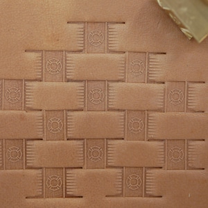 May include: Close-up of stamped leather with a repeating pattern of a firefighter emblem within a woven design. The leather is a light brown color, and a brass tool is visible in the upper right corner.