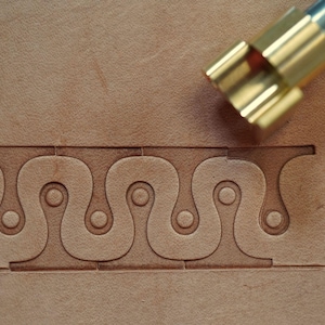 May include: Close-up of leather tooling with a decorative pattern. A brass tool is positioned above the leather, ready to create the design. The pattern features a repeating series of curved shapes and circles, creating a unique texture on the tan leather.