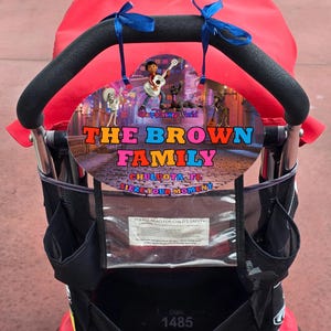 May include: A red and black stroller with a black handle and a decorative sign. The sign features characters from the movie "Coco" and the text "THE BROWN FAMILY, CHULUOTA, FL, SIEZE YOUR MOMENT".