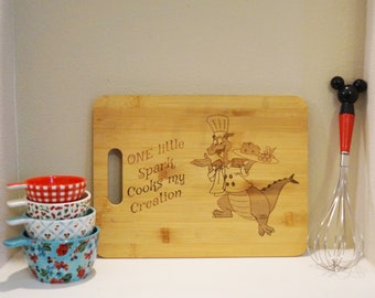 Puede incluir: Una tabla de cortar de madera con un chef dragón de dibujos animados sosteniendo un plato de comida. El texto "ONE little Spark Cooks my Creation" está grabado en la tabla.