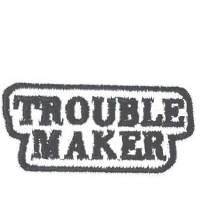 May include: A black and gray embroidered patch with the words "TROUBLE MAKER" in a bold, stylized font. The text is enclosed within a rectangular border with a fuzzy, textured appearance, set against a white background.