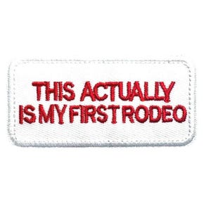 May include: Embroidered white patch with the red text: "THIS ACTUALLY IS MY FIRST RODEO." The patch has a rectangular shape with rounded corners and a stitched border. The text is in a bold, sans-serif font.