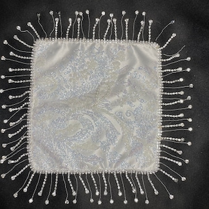 May include: A white square satin pillow with a silver glitter pattern and a fringe border of clear rhinestones.