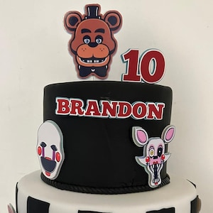 May include: A two-tiered black and white checkered cake decorated with cartoon characters from the video game Five Nights at Freddy's. The top tier is black with the name "BRANDON" and the number "10" in red. The bottom tier is white with black checkered squares. The cake is decorated with various characters, including Freddy Fazbear, Foxy the Pirate Fox, Chica the Chicken, and Bonnie the Bunny.