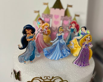 May include: A cake decorated with princess figurines and a pink castle backdrop. The cake is adorned with gold butterflies and a gold-framed pink oval with the name "Ayla" and the number "3".
