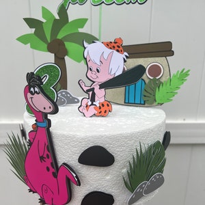 Puede incluir: Un adorno para pastel blanco con un personaje de dibujos animados sosteniendo un bate, un dinosaurio rosa, una palmera verde y las palabras "Ja'Seem".