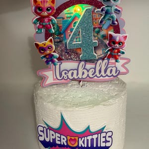 May include: A white cake decorated with a Super Kitties theme. The cake topper includes four cat figurines, the number 4, and the name "Isabella". The Super Kitties logo is displayed on the cake's front.