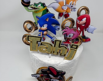 May include: A white cake decorated with gold glitter and a name "Tahi" in gold glitter. The cake is topped with six Sonic the Hedgehog characters: Sonic, Tails, Knuckles, Amy, Shadow, and Vector.