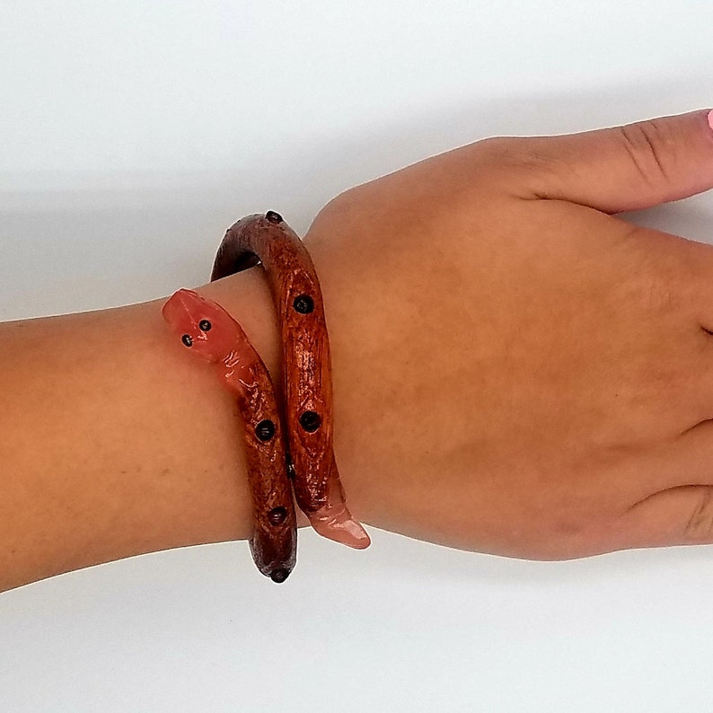 K&ouml;nnte beinhalten: Ein Holzschlangenarmband mit schwarzen Akzenten. Das Armband ist um das Handgelenk einer Person gewickelt.