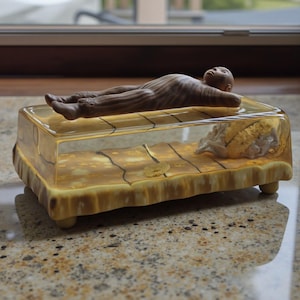 May include: A unique decorative piece featuring a small figure lying on a rectangular, transparent container. The container has a yellow-brown base with a textured design and a shell-like object inside. The figure is brown with detailed features.