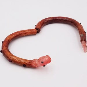K&ouml;nnte beinhalten: Ein Holz-Armband mit einem rosa Schlangenkopf-Anh&auml;nger und roten Edelstein-Akzenten.